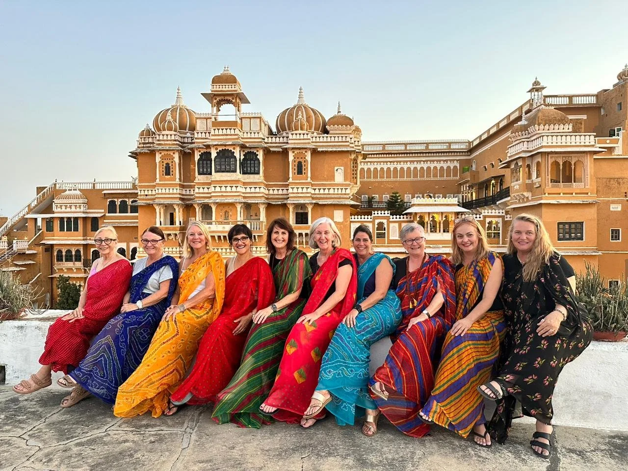 Ten women in bright saris seated on a wall in front of a Rajasthan palace